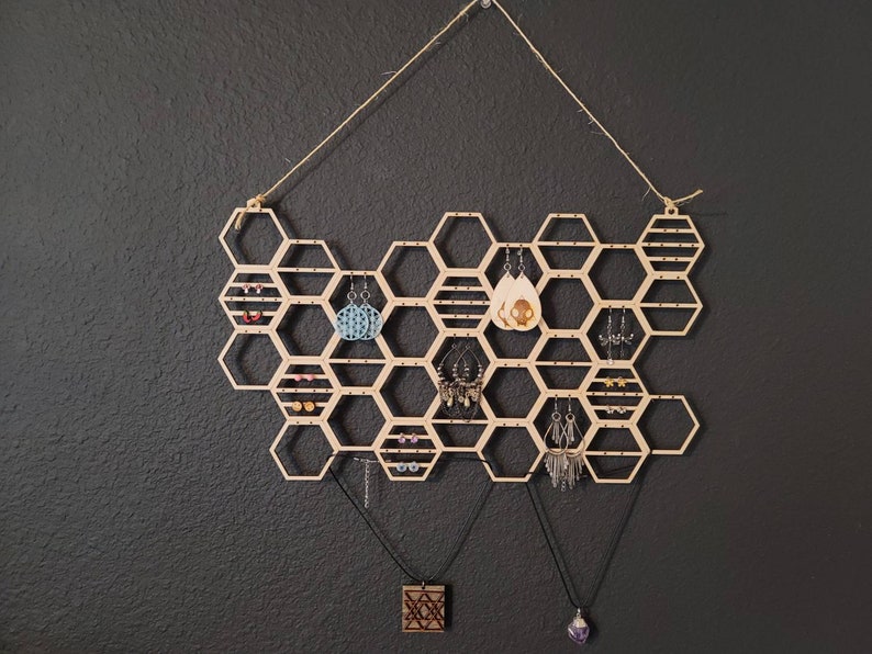 Puede incluir: Organizador de pendientes y collares de madera en forma de panal con m&uacute;ltiples compartimentos. El organizador est&aacute; colgado de una cuerda y tiene varios pendientes y collares colgando de &eacute;l.