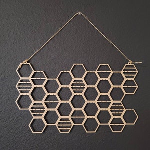 Puede incluir: Un colgante de pared de madera en forma de panal con una cuerda para colgar. El panal est&aacute; hecho de hex&aacute;gonos individuales con un patr&oacute;n de rayas.