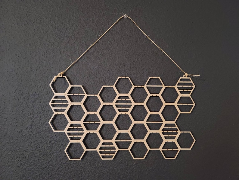 Puede incluir: Un colgante de pared de madera en forma de panal con una cuerda para colgar. El panal est&aacute; hecho de hex&aacute;gonos individuales con un patr&oacute;n de rayas.