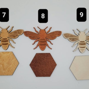 Puede incluir: Tres recortes de abejas de madera con diferentes manchas de madera. Las abejas son todas del mismo tama&ntilde;o y forma, con un cuerpo marr&oacute;n y rayas negras. Las abeilles est&aacute;n sentadas sobre posavasos hexagonales de madera. Los posavasos est&aacute;n te&ntilde;idos en madera natural, marr&oacute;n oscuro y marr&oacute;n claro.