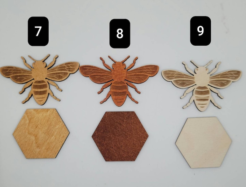 Puede incluir: Tres recortes de abejas de madera con diferentes manchas de madera. Las abejas son todas del mismo tama&ntilde;o y forma, con un cuerpo marr&oacute;n y rayas negras. Las abeilles est&aacute;n sentadas sobre posavasos hexagonales de madera. Los posavasos est&aacute;n te&ntilde;idos en madera natural, marr&oacute;n oscuro y marr&oacute;n claro.