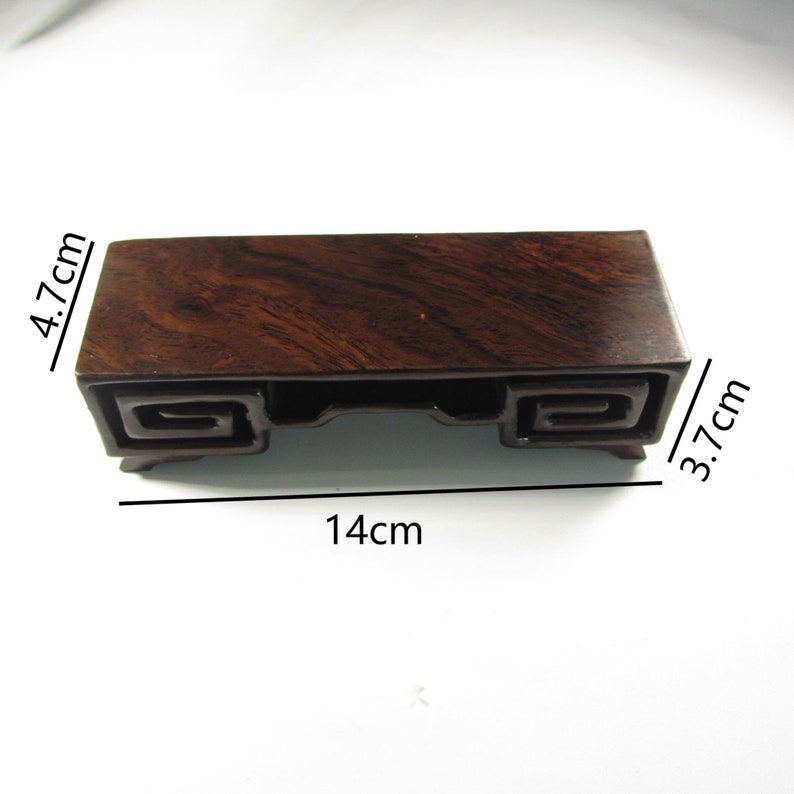 Op de afbeelding: Een donkerbruine houten displaystandaard met een rechthoekige vorm. De standaard is 14 cm lang, 4,7 cm hoog en 3,7 cm breed. Het heeft decoratieve krullen aan de zijkanten en is geschikt voor kleine objecten.