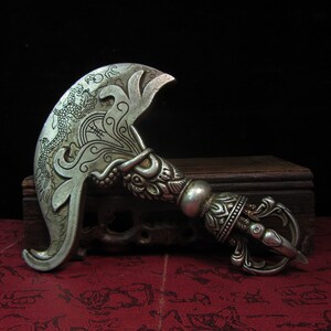 May include: Silver-colored ritual blade with intricate dragon and floral engravings. The handle features detailed carvings and a rounded pommel. The blade has a curved, crescent shape. The object is displayed on a wooden surface.