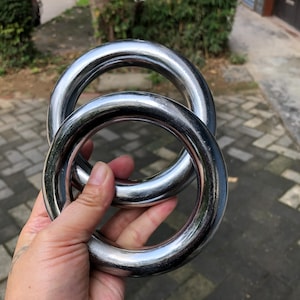 May include: Two large, shiny, silver metal rings are held in a hand. The rings are thick and circular, with a polished surface that reflects the surroundings. The rings are interlocked, suggesting they are a set or part of a larger design.