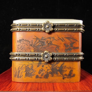 Puede incluir: Una caja rectangular de dos niveles con una tapa de color claro y un cuerpo marrón anaranjado cálido. La caja está adornada con intrincadas ilustraciones grabadas en marrón oscuro de aves y paisajes. Bandas metálicas decorativas con detalles florales rodean la caja.