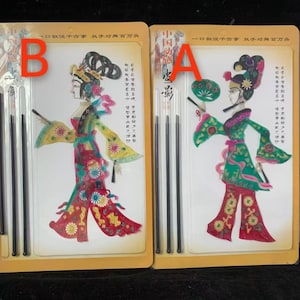 Puede incluir: Dos marionetas chinas de sombras empaquetadas. Cada marioneta es una figura colorida de una mujer con vestimenta tradicional, sosteniendo un abanico u otros accesorios. El embalaje incluye texto chino y largas varillas negras para la manipulación.