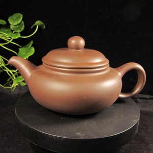 Può includere: Una teiera in ceramica marrone con corpo arrotondato, beccuccio curvo e manico ad anello. Il coperchio ha un piccolo pomello rotondo. La teiera è esposta su una superficie nera su sfondo scuro.