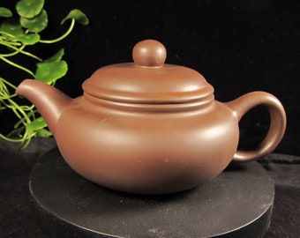 May include: A brown ceramic teapot with a rounded body, a curved spout, and a loop handle. The lid has a small, round knob. The teapot is displayed on a black surface against a dark background.