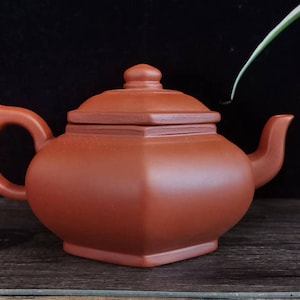 Peut inclure: Une théière en céramique brun-rouge avec une base et un couvercle carrés, une anse incurvée et un bec verseur. La théière a une finition lisse et mate et repose sur une surface en bois. Le design est simple et élégant, pour le thé.