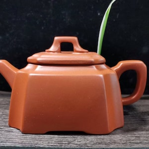 以下が含まれることがあります： 正方形のハンドルと蓋が付いた茶色のセラミックティーポット。 ティーポットは幾何学的なデザインで、注ぎ口が付いています。 ティーポットは木製の表面に置かれています。