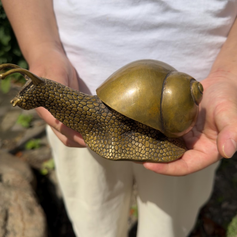 May include: A brass snail figurine featuring a highly textured, pebbled body and a smooth, polished, rounded shell. The metallic sculpture displays a warm, golden-brown patina with intricate detailing on the head, tentacles, and foot.
