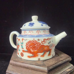 May include: Cream-colored ceramic teapot with a lid, decorated with a red and orange lion design. The lid features blue floral patterns and red dots. The teapot has a curved handle and a spout.