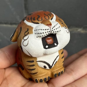 May include: A small, handcrafted ceramic tiger figurine. The tiger has a white face with black dots, brown and black stripes, and a smiling expression. The figurine is painted in shades of brown, tan, and white.
