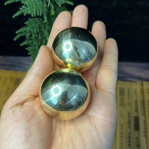 May include: Two polished, gold-colored metal spheres, each reflecting the surrounding environment. The spheres are held in a hand, with a dark background and a hint of greenery visible. The spheres appear to be smooth and seamless.