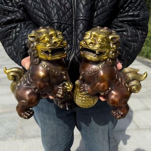 May include: Two bronze-colored Foo Dog statues with gold-colored accents. The statues are in a seated position, holding a ball. The Foo Dogs have detailed features, including open mouths and flowing manes. The statues are held up by a person.