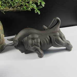 May include: A dark brown ceramic bull figurine, sculpted in a dynamic pose with its head lowered and tail raised. The bull has detailed musculature and curved horns, suggesting strength and power. This decorative piece is suitable for home decor.