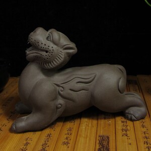 May include: A brown ceramic sculpture of a stylized lion in a reclining pose. The lion has detailed facial features, including an open mouth and textured mane. Decorative wing-like elements are on the sides. The sculpture is on a bamboo surface.
