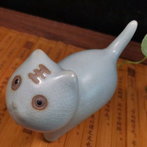 Może przedstawiać: Jasnoniebieska ceramiczna figurka kota z krakelą. Kot ma duże, okrągłe oczy i brązowe oznaczenie na czole. Figurka jest ustawiona z podniesionym ogonem.