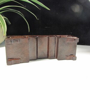 Op de afbeelding: Een donkerbruine, rechthoekige houten displaystandaard met verticale groeven en kleine vierkante uitsteeksels. Ontworpen om een klein object te bevatten, staat het op een wit oppervlak. Ideaal voor decoratie.