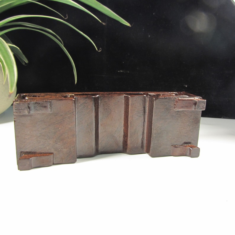 Op de afbeelding: Een donkerbruine, rechthoekige houten displaystandaard met verticale groeven en kleine vierkante uitsteeksels. Ontworpen om een klein object te bevatten, staat het op een wit oppervlak. Ideaal voor decoratie.