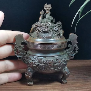 May include: Bronze incense burner with detailed dragon and cloud carvings. The burner features a dragon figure on the lid, two handles, and three feet. The design is ornate, showcasing traditional craftsmanship. The colour is a dark bronze.