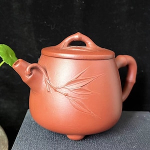 May include: A reddish-brown ceramic teapot with a lid and handle. The teapot features a bamboo leaf design carved on the side. The handle is curved, and the spout is angled. The teapot sits on a dark surface.