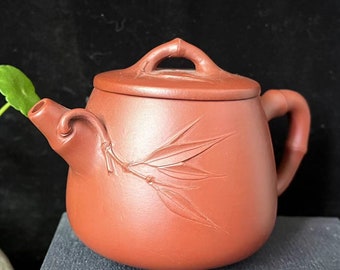 May include: A reddish-brown ceramic teapot with a lid and handle. The teapot features a bamboo leaf design carved on the side. The handle is curved, and the spout is angled. The teapot sits on a dark surface.