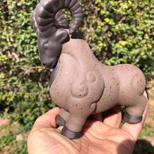 May include: A ceramic ram figurine in shades of brown and dark gray. The ram has large curved horns and a detailed body. The figurine is held in a hand, with a green leafy background.