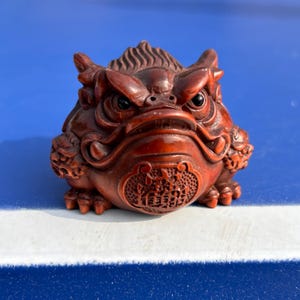 Puede incluir: Figurilla de rana tallada en color marrón rojizo con detalles intrincados. La rana tiene una expresión feroz, con ojos prominentes y una cresta detallada. La base presenta un patrón decorativo. La figurilla mide aproximadamente 10 cm de largo.