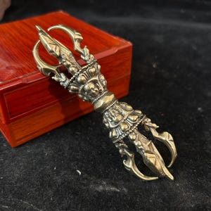 May include: A detailed brass vajra, a ritual object, with intricate carvings and a three-pronged design. The handle features detailed figures and a central grip. The vajra is set against a red wooden box and a dark background.