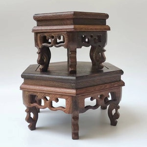 Puede incluir: Dos soportes de exhibición de madera marrón oscuro. El soporte superior es cuadrado y el inferior es hexagonal. Ambos soportes tienen patas talladas y adornos decorativos. La veta de la madera es visible y los soportes están apilados.