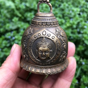 May include: A bronze-colored bell with a loop handle. The bell features a detailed design with floral patterns and a central medallion depicting a Buddha figure. The bell is held in a hand against a blurred green background.