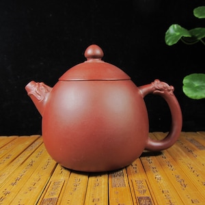 Puede incluir: Tetera de cerámica de color marrón rojizo con cuerpo redondeado, tapa con pequeño pomo y asa curvada. El pico y el asa están adornados con detalles de dragón. La tetera se exhibe sobre una superficie de bambú.