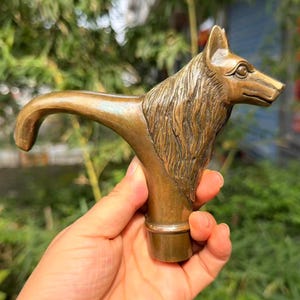 Op de afbeelding: Een metalen, bronskleurige wandelstokgreep met een gedetailleerde, gebeeldhouwde wolvenkop met gestructureerde vacht, puntige oren en een langwerpige snuit, uitlopend in een gebogen nekgedeelte dat eindigt in een cilindrische basis voor bevestiging.