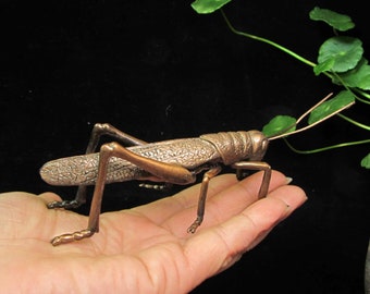 May include: A bronze grasshopper sculpture with detailed texture, held in a hand. The insect has long legs and antennae, with a dark background and green foliage in the upper right corner. The sculpture is a decorative item.