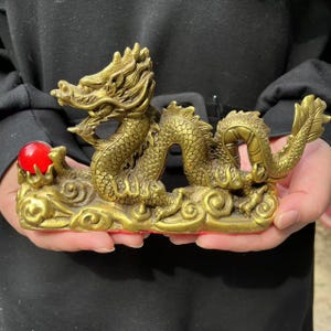 Peut inclure: Une sculpture de dragon en laiton doré avec un orbe rouge. Le dragon est posé sur une base en forme de nuage. La sculpture est de couleur dorée et présente des détails complexes.