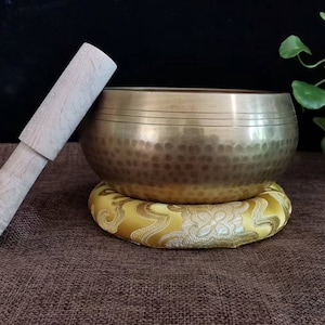 May include: A bronze singing bowl with a wooden mallet rests on a yellow floral cushion. The bowl has a textured surface and is positioned on a brown surface. The mallet is light-colored wood.