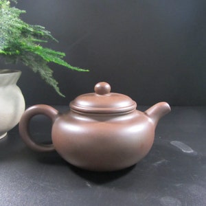 May include: A brown ceramic teapot with a rounded body, a curved handle, and a spout. The lid has a small knob on top. The teapot is on a black surface. A vase and greenery are in the background.