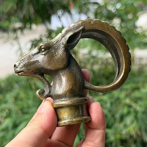 Puede incluir: Una escultura de metal en tono bronce de una cabeza de íbice con un cuerno grande y curvado que se dirige hacia el cuello. La pieza tiene una superficie texturizada y una base cilíndrica, mostrando rasgos faciales detallados y una barba.