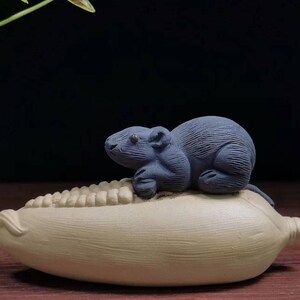 May include: A small, dark blue ceramic rat figurine perched on a tan corn cob. The rat has detailed features and is sitting upright. The corn cob has a textured surface. The background is dark.