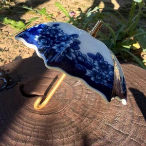 May include: A miniature umbrella-shaped ornament crafted from white ceramic with a deep blue floral pattern. The piece features a curved gold-toned metal handle and a small gold-toned finial at the top, creating a delicate and detailed decorative object.