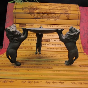 Könnte beinhalten: Eine bronzefarbene Skulptur mit drei Löwen, die eine flache, ovale Oberfläche tragen. Die Löwen stehen aufrecht, mit erhobenen Pfoten und Köpfen. Der Hintergrund zeigt Bambusrollen mit chinesischen Schriftzeichen.