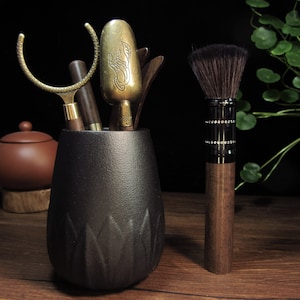 May include: A black ceramic tea tool holder with a set of tea ceremony tools and a makeup brush. The tools include a brass scoop with an etched design, wooden tools, and a brush with a dark wooden handle and black bristles.