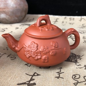 May include: A small, reddish-brown ceramic teapot with a lid and handle. The teapot is decorated with embossed floral designs. The spout and handle are curved. The teapot is sitting on a patterned surface.