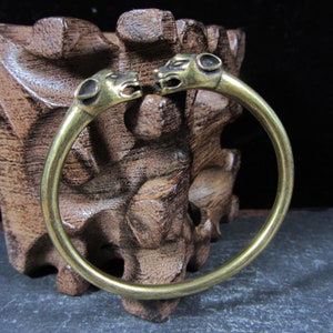 May include: A brass-colored bangle bracelet featuring two animal head sculptures facing each other. The bracelet has a circular design and is made of a shiny, metallic material. The animal heads have detailed features, including open mouths and visible ears.