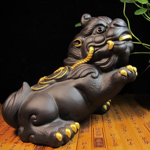 May include: A dark brown ceramic sculpture of a mythical creature with gold accents. The creature has a lion-like body, a fierce expression, and is posed in a seated position. The sculpture features intricate details and is likely a decorative item.