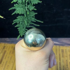 May include: A polished, metallic sphere held in a hand. The sphere reflects the surrounding environment, including a circular light and greenery. The surface of the sphere is smooth and reflective, with a golden hue at the base.
