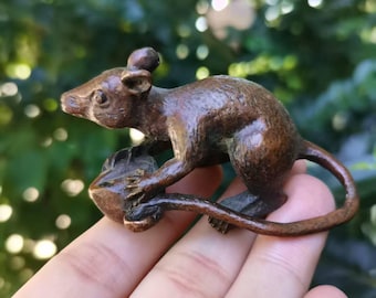 May include: A bronze-colored rat figurine, holding a small object. The rat is detailed with visible ears, whiskers, and a long tail. The sculpture is held in a hand, with a blurred green background.