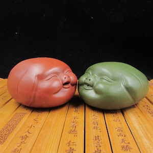 Könnte beinhalten: Zwei Keramik-Schweinefiguren, eine rote und eine grüne, mit lächelnden Gesichtern. Die Figuren sind glatt und abgerundet, mit detaillierten Gesichtszügen. Sie stehen auf einer Holzoberfläche vor einem dunklen Hintergrund.