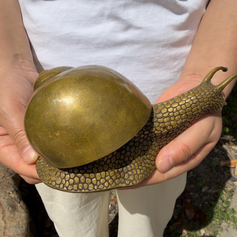 May include: A brass snail figurine featuring a smooth, polished, rounded shell and a textured body with a detailed, pebbled surface pattern. The sculpture has two prominent eye stalks extending from the head and a curved, elongated neck.
